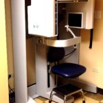 A specialized panoramic dental X-ray machine (CBCT) located in a dedicated imaging room with checkered flooring.